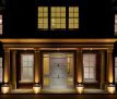 a stunning entrance to a luxury mansion taken at night with interior and external lights switched on. A large porch supported by six fluted Doric columns is beautifully lit from ground and ceiling lights. The subtle differences in colour from the various Georgian windows reflects the temperature of the wall coverings and internal lights. A large white solid wood front door surrounded by glass panels sits in the centre.