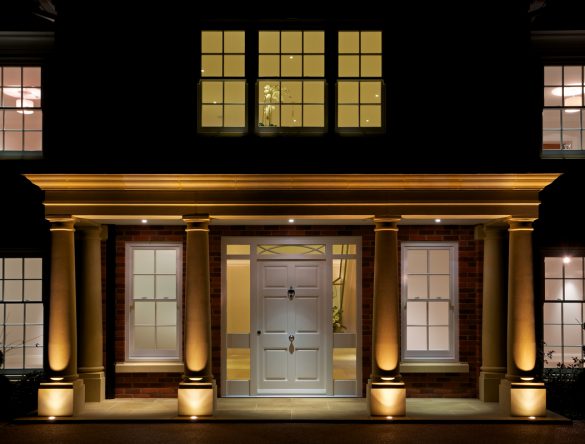 a stunning entrance to a luxury mansion taken at night with interior and external lights switched on. A large porch supported by six fluted Doric columns is beautifully lit from ground and ceiling lights. The subtle differences in colour from the various Georgian windows reflects the temperature of the wall coverings and internal lights. A large white solid wood front door surrounded by glass panels sits in the centre.